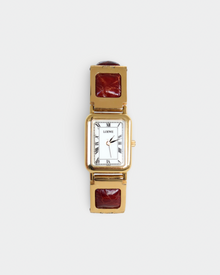 A women's rectangular analog watch with a gold-tone case, white dial with black Roman numerals, and a gold-tone link bracelet featuring square dark red inlays, shown on a light gray background.
