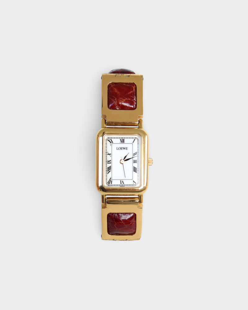 A women's rectangular analog watch with a gold-tone case, white dial with black Roman numerals, and a gold-tone link bracelet featuring square dark red inlays, shown on a light gray background.